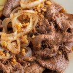 A serving of Pinoy Beef Bistek Recipe with thin slices of beef, sautéed onion rings, and golden potato wedges on white rice, styled on a plate for a Filipino comfort food meal.